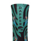 Back view of a black seamless toe sock with emerald green swirls and blue and yellow parrot design print and with a emerald green strip with Dapper Chameleon wording and logo in black.